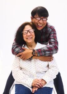Allen Galeon, a Filipino care worker and family caregiver, is leaning over and hugging his mother, Pura Sherri Bloom, a former home care worker, who is seated in front of him. Allen is wearing a red and grey plaid shirt, glasses, and black pants, and Pura is wearing a white polka dot shirt and jeans. 