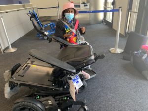 Engracia, in the background, seated in a temporary wheelchair, faces the camera and is wearing a pink hat and mask. In front of her is her broken power wheelchair, with the backrest completely bent back. 